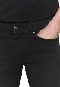 Jeans de denim negro con un corte slim, con cierre de botones, bolsillos delanteros y una textura suave. Detalles de costuras visibles a lo largo de los bordes.
