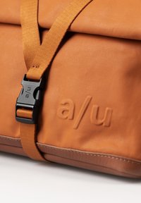 Orangefarbene Tasche mit matter Textur, ausgestattet mit einer schwarzen Kunststoffschnalle und geprägtem Logo. Sie verfügt über einen breiten Schulterriemen und einen kontrastierenden unteren Abschnitt.