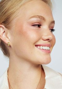 Guldörhänge som visas på en ljushyad modell. Örhänget har en slät, glänsande yta och en modern, rundad design.