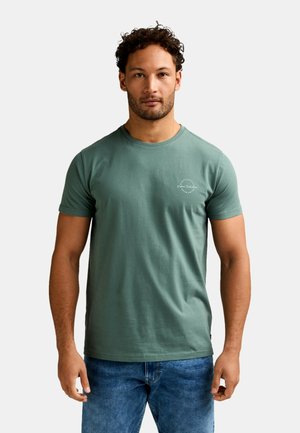 Uomo con capelli corti e ricci e barba chiara, che indossa una maglietta verde a maniche corte e jeans blu, in piedi contro uno sfondo chiaro uniforme.
