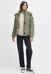 Olivengrønn pufferjakke med høy krage, polstret design og svart tilbehør. Båret over en beige strikkegenser og svarte bukser, sammen med sneakers.