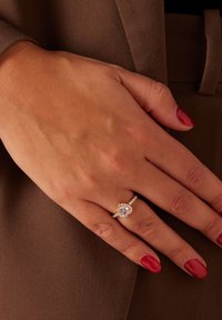 Gouden verlovingsring met een hartvormige centrale steen, omringd door kleinere diamanten op een gestructureerde band. Hand rust op een bruine ondergrond.
