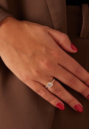 Gouden verlovingsring met een hartvormige centrale steen, omringd door kleinere diamanten op een gestructureerde band. Hand rust op een bruine ondergrond.