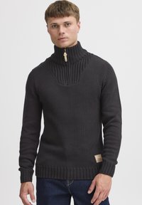 Pull en tricot noir avec un col côtelé haut et une ouverture zippée, présentant des motifs texturés et un patch logo en bas à droite.