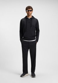 Uomo in piedi con le mani nelle tasche, indossando una felpa nera con cappuccio, pantaloni coordinati, sneakers sportive e occhiali da sole scuri a mascherina.