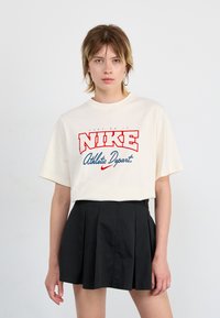Crème-kleurige katoenen T-shirt met korte mouwen en een ronde halslijn, met een gedurfde rode en blauwe Nike-afbeelding op de voorkant.
