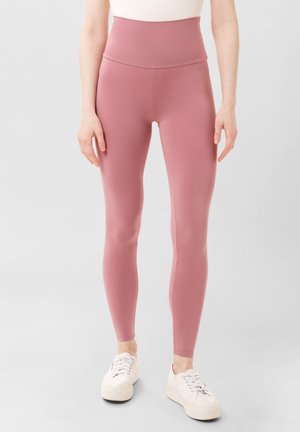 Leggings a vita alta, aderenti, in tessuto rosa morbido con design senza cuciture. Abbinati a scarpe da ginnastica bianche, le gambe sono affusolate alla caviglia.