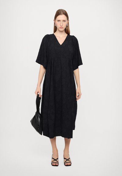 Zwarte midi-jurk met korte mouwen, V-hals, textuurstof en een losse fit. Gepaard met een zwarte tas en neutrale sandaaltjes met bandjes.