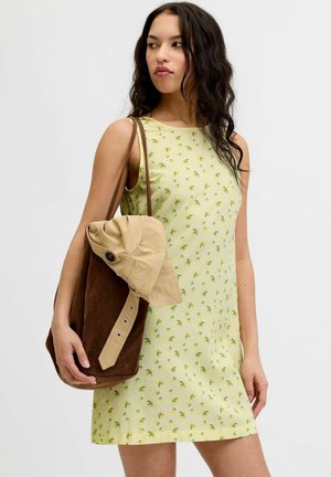 Mujer con vestido amarillo sin mangas con pequeño estampado floral, sosteniendo un bolso tote marrón con un sombrero beige colocado encima.