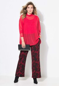 Rotes Netzoberteil mit hohem Ausschnitt und Verzierungen, kombiniert mit schwarzen und roten gemusterten weit geschnittenen Hosen. Schwarze Clutch mit weißen Akzenten.