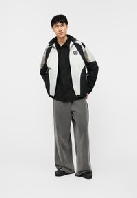 Νεαρός άνδρας στέκεται φορώντας μαύρο και λευκό μπουφάν με φερμουάρ, μαύρο πουκάμισο με ρίγες, φαρδιά γκρι παντελόνια και μαύρα παπούτσια μπροστά σε απλό φόντο.