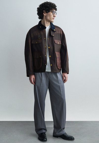 Jeune homme aux cheveux bouclés et portant des lunettes, vêtu d'une veste marron, d'une chemise blanche, d'un pantalon gris à jambes larges et de chaussures noires, debout de profil.