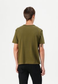 Olivenfarbendes Baumwoll-T-Shirt mit kurzen Ärmeln, Rundhalsausschnitt und geradem Saum. Auf dem rechten Arm ist ein kleines Tattoo sichtbar.