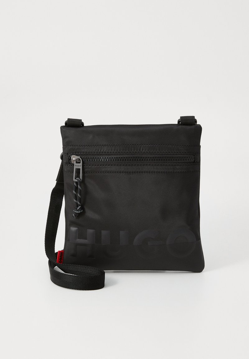 Sacolina transversal en nylon noir avec une bandoulière réglable, poche zippée à l'avant et logo en relief. Texture lisse et design minimaliste.