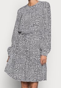 Femme portant une robe à manches longues en motif pied-de-poule noir et blanc, avec une taille cintrée et une jupe fluide.