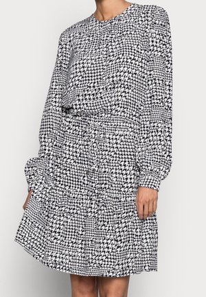 Femme portant une robe à manches longues en motif pied-de-poule noir et blanc, avec une taille cintrée et une jupe fluide.