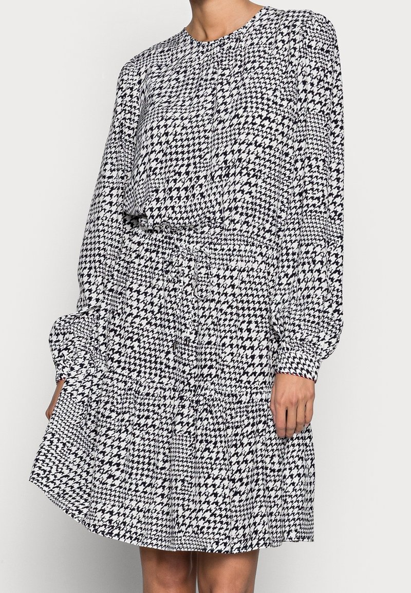 Femme portant une robe à manches longues en motif pied-de-poule noir et blanc, avec une taille cintrée et une jupe fluide.