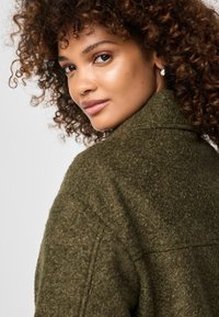 Een vrouw met krullend haar draagt een gestructureerde, olijfgroene wollen jas met een kraag, die subtiele variaties in de oppervlakte van de stof toont.