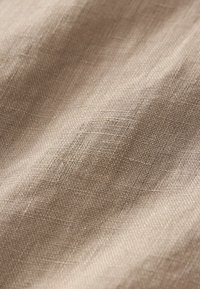 Beige geweven stof met een textuur en een licht gekreukeld oppervlak, waarbij gedetailleerde linnen-achtige vezels en kruispatronen zichtbaar zijn.