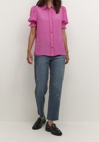 Blusa fucsia de manga corta con cuello, mangas abullonadas y cierre de botones al frente. Combinada con jeans rectos azules y mocasines negros.