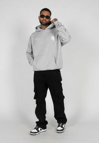 Sudadera con capucha gris y bolsillo frontal, pantalones cargo negros con bolsillos laterales y zapatillas negras y blancas. Gafas de sol negras como accesorio.