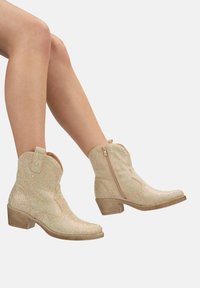 Botas de vaquero beige brillantes con punta afilada, tacón apilado y cremallera lateral, que presentan acentos brillantes en toda la superficie y detalles cosidos.