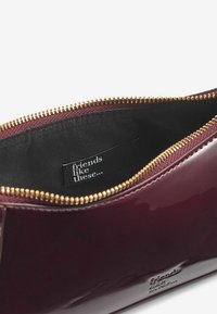 Burgunderfarbene Lackleder-Reißverschluss-Tasche mit einem goldenen Reißverschluss, ausgestattet mit einem schwarzen Innenetikett, auf dem steht "Freunde wie diese..."