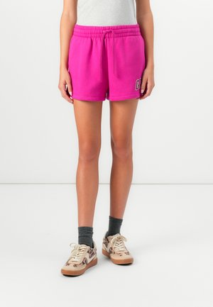 Shorts en coton rose avec une taille élastique et un cordon de serrage, présentant un logo imprimé sur la jambe gauche. Portés avec des baskets à motifs et des chaussettes grises.