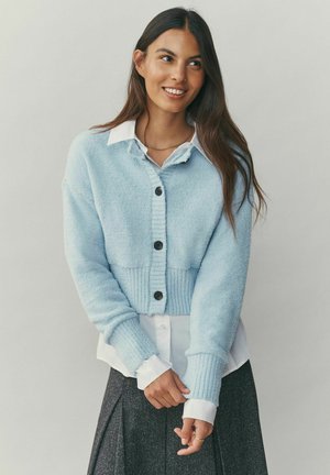 Vaaleansininen lyhyt neuletakki, valmistettu teksturoidusta kankaasta, jossa on resorivyötärö, viisi mustaa nappia ja pitkät hihat valkoisen paidan päällä.