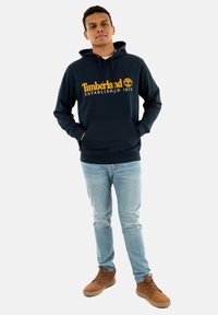 Sudadera de marina con el texto amarillo "Timberland Established 1973", bolsillo tipo canguro y dobladillo acanalado. Combinada con jeans azul claro y botas marrones.