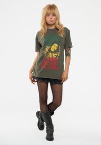 Olive cotton t-shirt featuring a colorful graphic of a person with the text "One Love." Worn with a denim skirt and black knee-high boots.