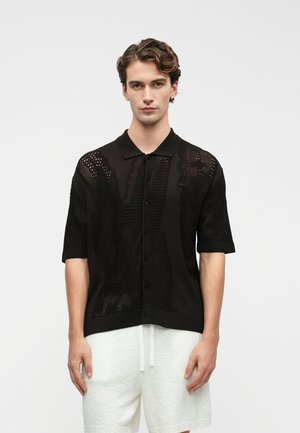 Polo noir à manches courtes avec un design en maille ouverte texturée, patte de boutonnage et col et poignets côtelés. Esthétique simple et moderne.