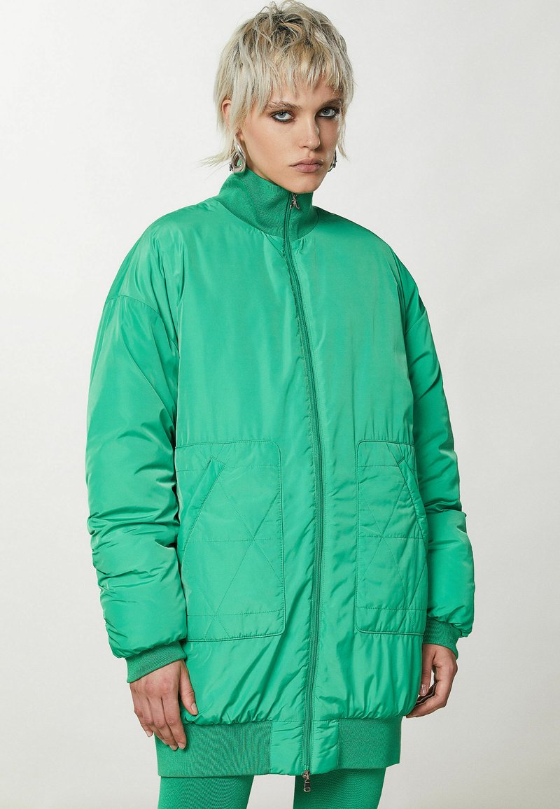 Zielona pikowana kurtka z wysokim kołnierzem, zamkiem błyskawicznym z przodu i dwoma kieszeniami z przodu. Posiada ściągacze przy mankietach i luźny krój. Gładka faktura.