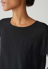 Zwarte, kortemouwen top met een ronde halslijn. Glad, licht materiaal met een subtiele naaddetail over de borst. Geen zichtbare patronen.