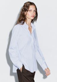 Camicetta azzurra chiara con colletto a punta, maniche lunghe e spacchi laterali. Indossata con pantaloni marroni scuri, evidenziando una vestibilità rilassata.