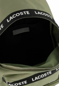 Mochila de tela verde con un forro interior negro. Presenta un logo blanco de Lacoste en la correa y un cierre de cremallera. Diseño simple y funcional.