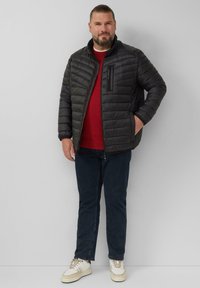 Zwarte gewatteerde pufferjack met een hoge kraag en een ritszak op de borst, gedragen over een rode trui, in combinatie met donkere jeans en witte sneakers.