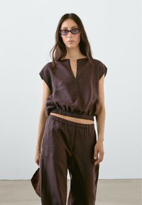 Linen cropped top with a V-neck and elastic waist, paired with loose-fitting pants in dark brown. Accessories include rectangular sunglasses and a bag.