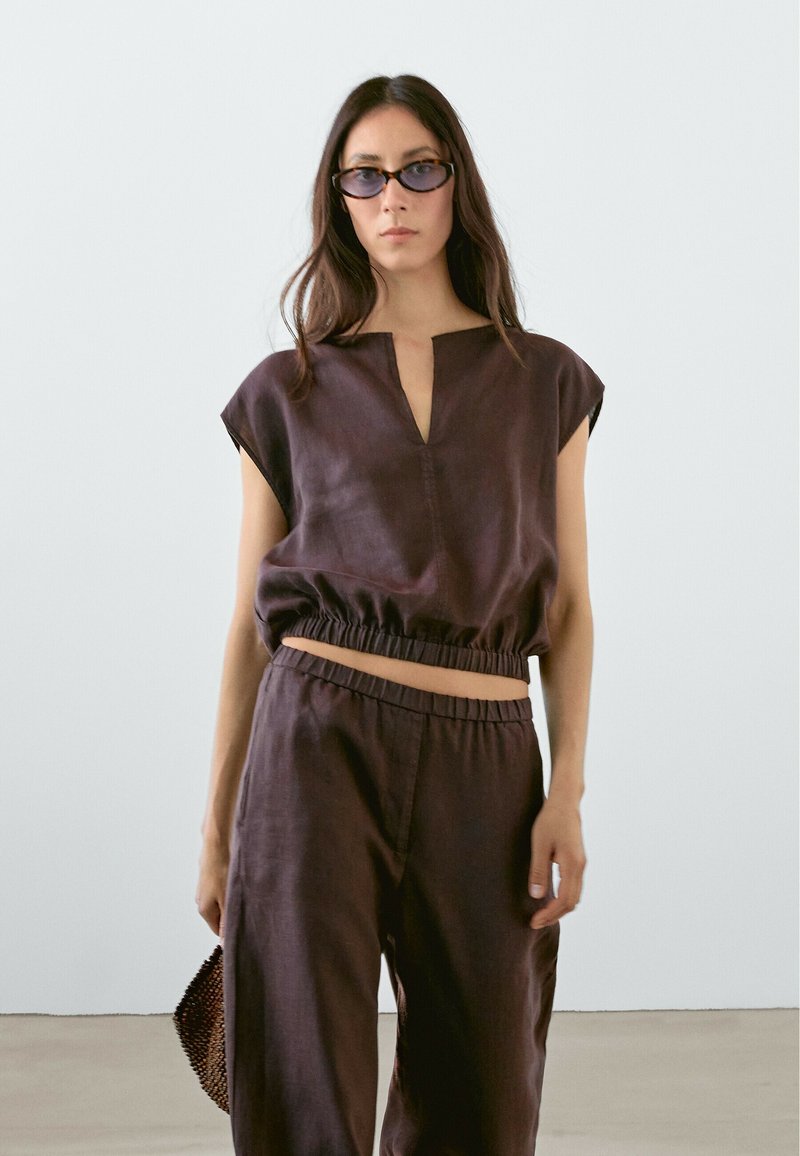 Linen cropped top with a V-neck and elastic waist, paired with loose-fitting pants in dark brown. Accessories include rectangular sunglasses and a bag.