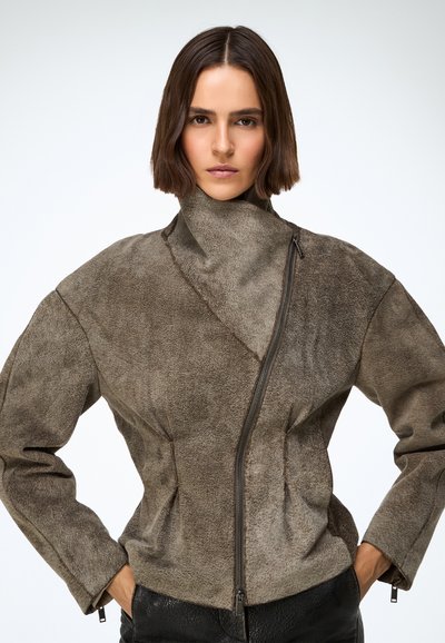 Barna velúr dzseki magas gallérral, aszimmetrikus cipzárral és oldalt ráncolt részletekkel, hosszú ujjakkal és texturált felülettel.