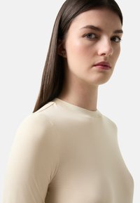 Beige top s dlouhým rukávem a vysokým výstřihem, hladká textura, přiléhavý střih a minimální švy. Část modelovy tváře je viditelná.
