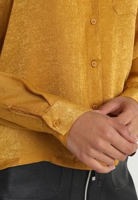 Chemise jaune moutarde, à boutonnage frontal, avec un tissu texturé et des manches longues. Présente une poche poitrine et des poignets à boutons.