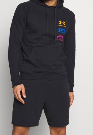 Man wearing black Under Armour hoodie with colorful logos and matching black shorts, standing against light background.