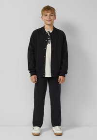 Lächelnder Junge steht vor einer grauen Wand, trägt eine schwarze knöpfbare Jacke, ein weißes T-Shirt mit Druck, schwarze Hosen und weiße Sneaker.