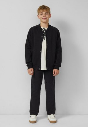 Uśmiechnięty chłopiec stoi przy szarej ścianie, ubrany w czarną zapinaną kurtkę, białą koszulkę z nadrukiem, czarne spodnie i białe sneakersy.