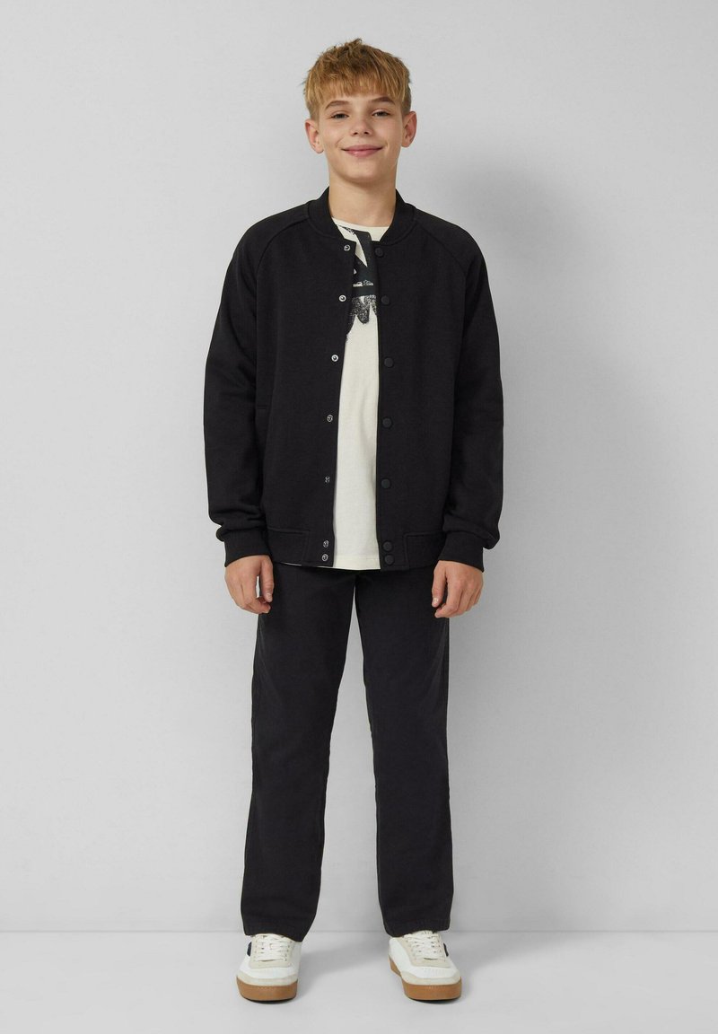 Lächelnder Junge steht vor einer grauen Wand, trägt eine schwarze knöpfbare Jacke, ein weißes T-Shirt mit Druck, schwarze Hosen und weiße Sneaker.