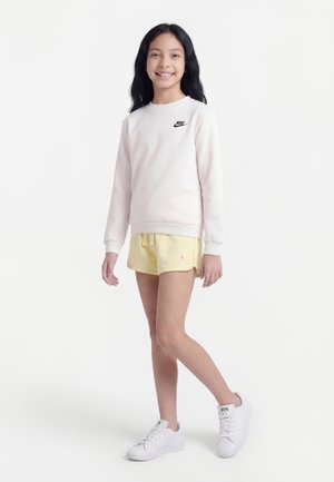 Niña joven de pie, vistiendo un sudadera blanca de Nike, pantalones cortos deportivos amarillo pálido y zapatillas blancas, sonriendo a la cámara sobre un fondo blanco.