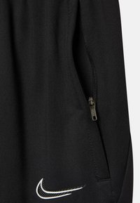 Pantalons de sport noirs avec une poche latérale zippée, texture lisse et un accent du logo Nike blanc. Le matériau semble respirant et extensible.