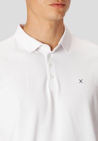 Hvid polo shirt med et struktureret mønster, to-knap lukning og et lille marineblåt logo på venstre bryst. Blødt bomuldsblandingsmateriale.