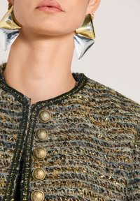 Nahaufnahme einer Person, die geometrische Ohrringe aus Gold und Silber sowie eine strukturierte gewebte Jacke mit dekorativen goldenen Knöpfen trägt.